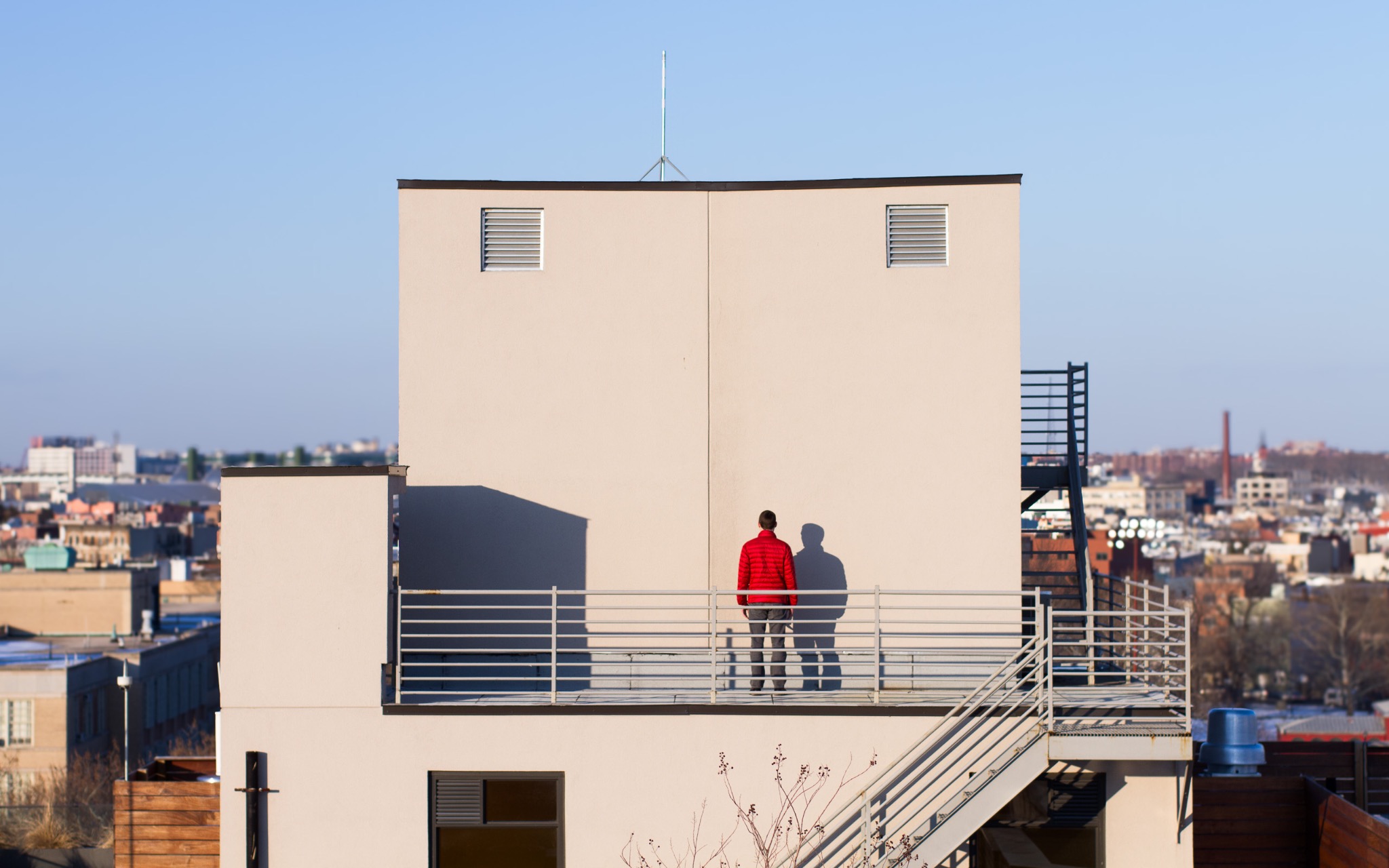 A lone figure in a red jacket on a sunlit rooftop beneath a blue sky